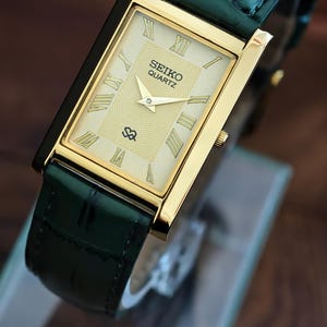 May include: A rectangular gold-toned Seiko Quartz watch with a cream-coloured face and Roman numeral hour markers. The watch has a dark green leather strap. The watch face displays the Seiko Quartz logo. The watch is displayed on a clear surface.