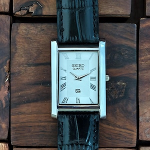 May include: A rectangular Seiko Quartz watch with a silver case and a white face featuring Roman numerals and the brand name. The watch has a black leather band with a crocodile pattern.