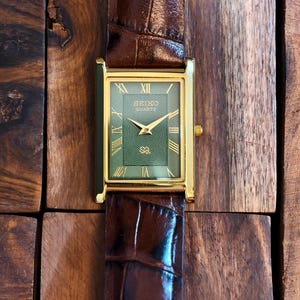 May include: A rectangular gold-toned Seiko Quartz watch with a green face and gold hands and Roman numerals. The watch has a brown leather strap with a crocodile pattern. The watch is displayed on a wooden surface.