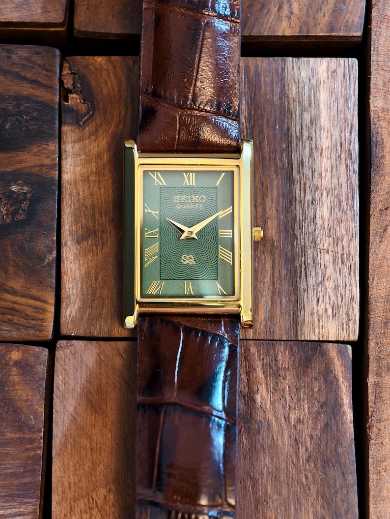May include: A rectangular gold-toned Seiko Quartz watch with a green face and gold hands and Roman numerals. The watch has a brown leather strap with a crocodile pattern. The watch is displayed on a wooden surface.