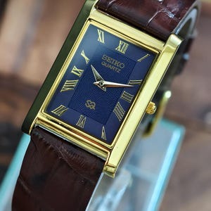 Op de afbeelding: Een rechthoekig Seiko Quartz horloge met een goudkleurige kast en een donkerblauwe wijzerplaat. Het horloge heeft gouden Romeinse cijfers en wijzers, en een bruine leren band. Een klassiek horloge.