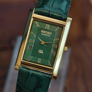 May include: A rectangular Seiko Quartz watch with a green face and gold-tone case. The watch features Roman numeral hour markers and a green leather strap. The watch face has the Seiko Quartz logo and a textured design.