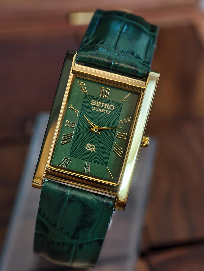 May include: A rectangular Seiko Quartz watch with a green face and gold-tone case. The watch features Roman numeral hour markers and a green leather strap. The watch face has the Seiko Quartz logo and a textured design.