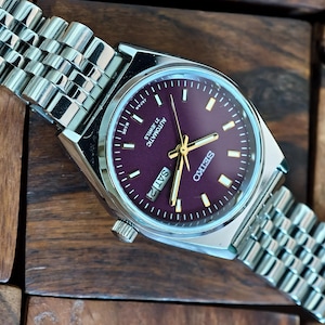 May include: A silver-toned Seiko automatic wristwatch with a burgundy dial and gold hands. The watch features a day-date display and a stainless steel band. The watch is displayed on a wooden surface.