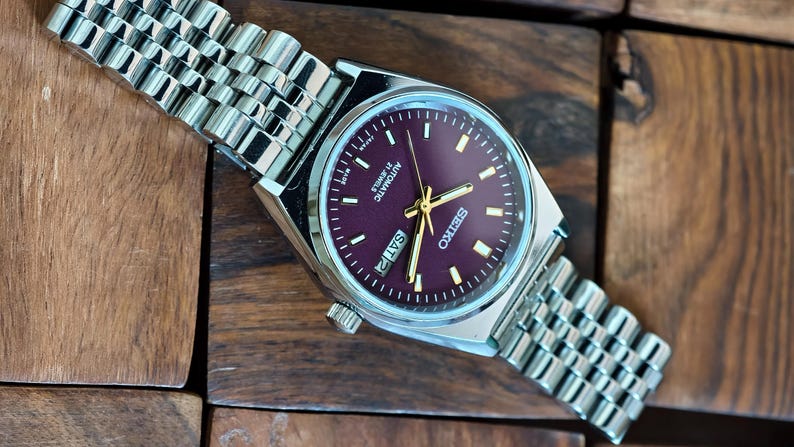 May include: A silver-toned Seiko automatic wristwatch with a burgundy dial and gold hands. The watch features a day-date display and a stainless steel band. The watch is displayed on a wooden surface.
