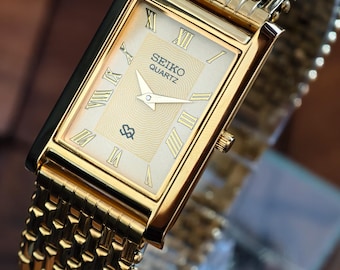 May include: A gold-toned Seiko Quartz watch with a rectangular face and a matching woven metal band. The watch face features Roman numerals and the Seiko logo. The watch is displayed on a glass surface.