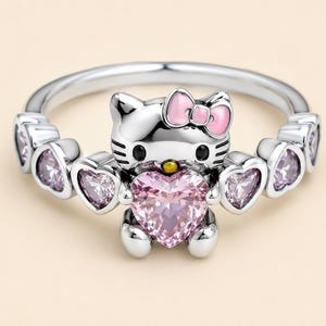 May include: A silver-toned ring featuring a cat character with a pink enamel bow, holding a large heart-shaped pink gemstone. The band is decorated with smaller heart-shaped settings containing light purple stones on both sides of the center.