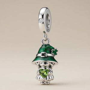 May include: A sterling silver charm bead featuring a figure wearing a green glittery hat with a clover accent and a black band. The figure holds a heart-shaped green gemstone. The bail is stamped with S925 and includes a small clear stone.
