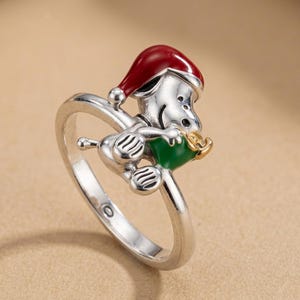 Peut inclure: Une bague en métal argenté représentant un personnage de chien de dessin animé portant un bonnet de Père Noël en émail rouge et tenant un petit cadeau vert avec un nœud doré. L'anneau est lisse et poli, avec un design tridimensionnel.