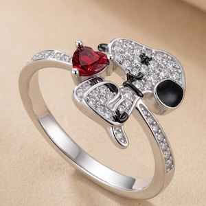 Puede incluir: Un anillo plateado con un diseño de personaje de dibujos animados. El anillo tiene una gema roja en forma de corazón y está adornado con pequeñas gemas transparentes. El personaje tiene detalles en negro y un ojo negro.