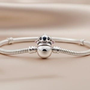 May include: A silver bracelet featuring a charm of a cartoon character with black eyes and pink ears. The bracelet has a snake chain design and is displayed on a beige satin surface. The charm is centered on the bracelet.