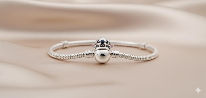 May include: A silver bracelet featuring a charm of a cartoon character with black eyes and pink ears. The bracelet has a snake chain design and is displayed on a beige satin surface. The charm is centered on the bracelet.