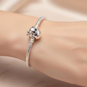 May include: A silver bracelet with a charm shaped like a dog's head. The charm has black eyes and pink accents. The bracelet is on a wrist, with a soft, neutral background.