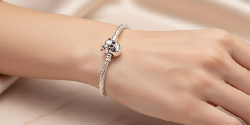 May include: A silver bracelet with a charm shaped like a dog's head. The charm has black eyes and pink accents. The bracelet is on a wrist, with a soft, neutral background.
