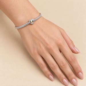 May include: A silver snake chain bracelet featuring a heart-shaped charm with a small Snoopy character graphic. The charm is centered on the metallic band, which has a textured, flexible design that wraps around the wrist with a polished finish.