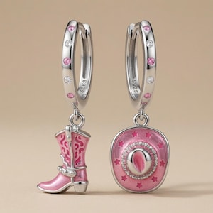 May include: A pair of silver hoop earrings featuring S925 hallmarks. One earring has a pink enamel cowboy boot charm, and the other has a pink enamel cowboy hat charm. Both hoops are decorated with alternating small pink and clear round stones.