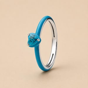 Puede incluir: Un anillo de plata con una banda de esmalte azul brillante y un engaste de piedra turquesa en forma de corazón. La piedra muestra un patrón natural de vetas negras y está asegurada por tres pequeñas garras de plata. El interior es de plata pulida.