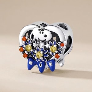 Peut inclure: Un charm en forme de cœur en argent représentant le personnage Snoopy en blanc sur le dessus. Le motif comprend des fleurs bleues aux centres jaunes, des accents orange et des détails en émail bleu à la base. La surface est polie et brillante.