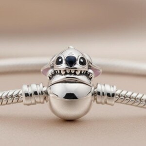 May include: A silver bracelet featuring a charm of a cartoon character with black eyes, a wide grin, and pointed ears. The bracelet has a snake chain design and is displayed against a soft, neutral background.