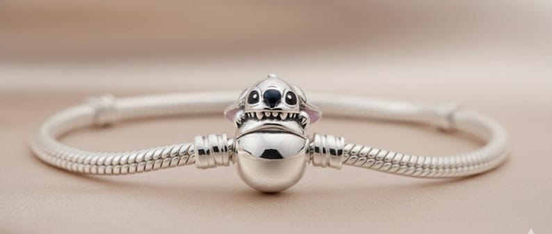May include: A silver bracelet featuring a charm of a cartoon character with black eyes, a wide grin, and pointed ears. The bracelet has a snake chain design and is displayed against a soft, neutral background.