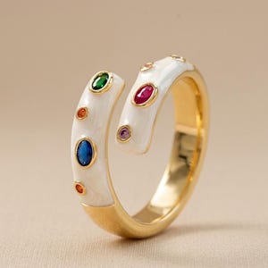 Puede incluir: Un anillo dorado con una banda de esmalte blanco, adornado con piedras preciosas de colores. El anillo presenta un diseño abierto, con un espacio entre los dos extremos. Las piedras preciosas son de varios colores, incluyendo verde, azul, rojo y naranja.