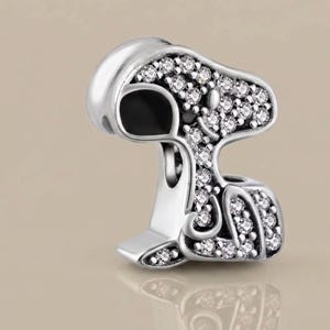 Peut inclure: Un charm en métal argenté en forme de chien de dessin animé, doté d'une oreille et d'un nez en émail noir lisse, tandis que le reste du corps est incrusté de nombreuses petites pierres rondes, claires et scintillantes.