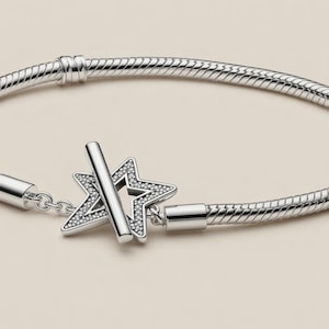 May include: A silver-toned snake chain bracelet featuring a toggle clasp with a star-shaped charm. The star charm is encrusted with small, clear, sparkling stones. The bracelet includes two fixed stations along the chain and a polished metal finish.