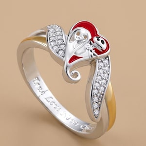 Puede incluir: Un anillo de tono plateado con una pieza central en forma de corazón, esmalte rojo y un diseño de personaje. La banda está decorada con pequeñas piedras transparentes y secciones doradas. El interior tiene grabado True Love Never Dies.