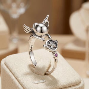 May include: A silver open ring featuring a winged koala figure reaching towards a circular charm with a star emblem. The band is stamped with S925 and includes a small, round, magenta-coloured gemstone set near the star charm on the ring band.
