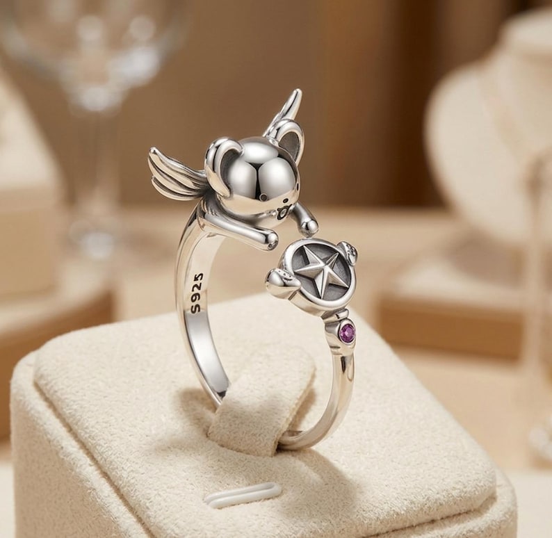 May include: A silver open ring featuring a winged koala figure reaching towards a circular charm with a star emblem. The band is stamped with S925 and includes a small, round, magenta-coloured gemstone set near the star charm on the ring band.