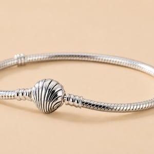 Puede incluir: Una pulsera de cadena de serpiente en tono plateado con un cierre central en forma de concha marina. La pulsera tiene una textura metálica lisa y flexible con un acabado pulido y un mecanismo de cierre redondeado inspirado en una concha.
