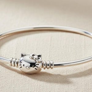 Pode incluir: Uma pulseira de prata com um berloque da Hello Kitty. O berloque apresenta a famosa personagem com um laço. A pulseira é feita de metal polido e tem um design simples e elegante. Circunferência de aproximadamente 18 cm.