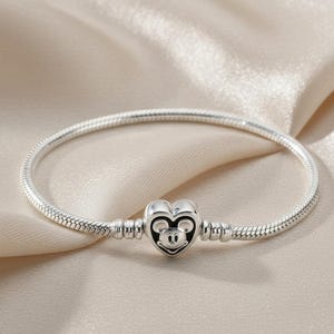 Pode incluir: Uma pulseira de prata com um berloque em forma de coração com um design do Mickey Mouse. A pulseira tem um design de corrente de cobra e é exibida em um tecido creme.