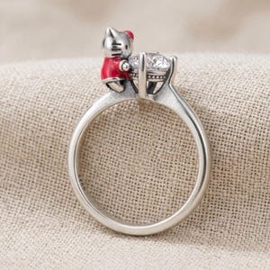 Puede incluir: Un anillo plateado con una figura de gato de dibujos animados con un atuendo rojo, colocado junto a una gran gema transparente. El anillo tiene una banda simple y un engaste detallado para la gema, sobre un fondo de tela neutra.