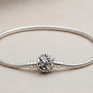 Puede incluir: Una pulsera plateada con un diseño de cadena de serpiente. La pulsera presenta un charm decorativo con detalles de corona y estrellas. La pulsera se muestra sobre un fondo neutro.