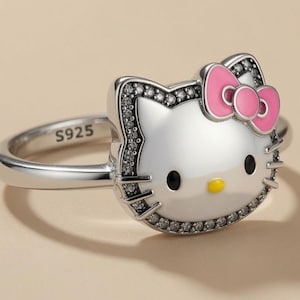May include: A silver ring featuring a white cat face with black eyes, a yellow nose, and a pink bow. The cat face is outlined with small clear stones, and the inner band is stamped with S925. The design is polished and metallic in appearance.
