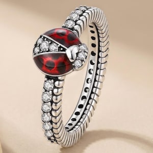 May include: A silver ring featuring a ladybug motif with a red enamel body and black spots. The band is textured with a ribbed pattern and is accented with clear round stones along the shank and around the ladybug's head and wings.