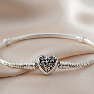 Puede incluir: Una pulsera de cadena de serpiente plateada con un cierre en forma de corazón. El cierre presenta un diseño en relieve oxidado de un árbol con ramas y hojas extendidas, creando un contraste detallado sobre el metal plateado pulido.