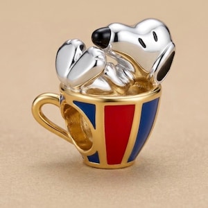 Peut inclure: Un charm argenté représentant un chien de dessin animé assis dans une tasse à thé dorée. La tasse présente des panneaux verticaux en émail rouge et bleu. Le chien a un nez noir et ses pattes sont levées près de sa tête.