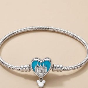 May include: A silver snake chain bracelet featuring a heart-shaped charm with a bright blue enamel background and a silver castle illustration. A small white Mickey Mouse head silhouette charm hangs from the bottom of the heart-shaped centerpiece.