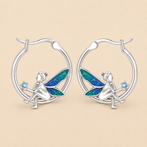 Puede incluir: Un par de pendientes de aro de plata con una figura sentada con alas brillantes azules y verdes. Cada pendiente muestra una pequeña piedra azul claro sostenida por la figura. El metal tiene el sello S925 de plata de ley.