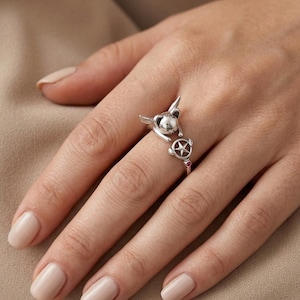 May include: A silver-toned open ring featuring a small creature with pointed ears and a circular charm containing a star, accented with a tiny pink stone on the band. The design wraps around the finger with the creature and star facing each other.