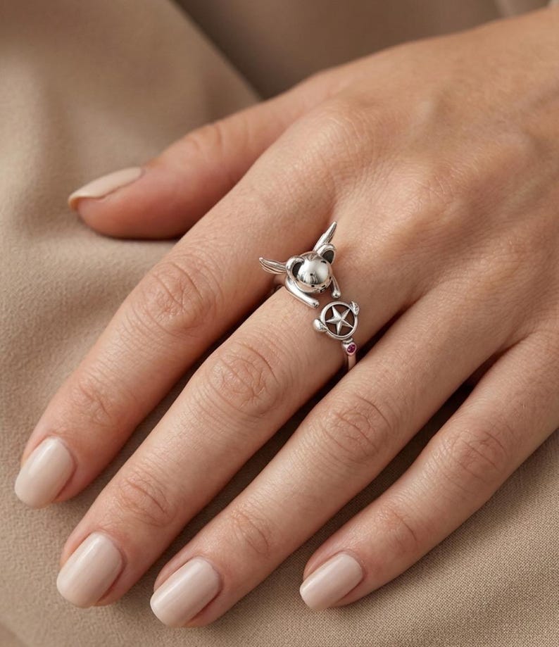 May include: A silver-toned open ring featuring a small creature with pointed ears and a circular charm containing a star, accented with a tiny pink stone on the band. The design wraps around the finger with the creature and star facing each other.