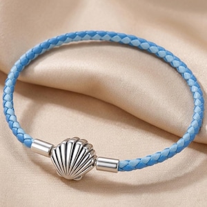 Puede incluir: Una pulsera de cuero trenzado azul con un cierre de concha plateada. La pulsera presenta un diseño azul de dos tonos, con un azul más claro tejido en el cuero azul más oscuro. El cierre de concha es detallado.