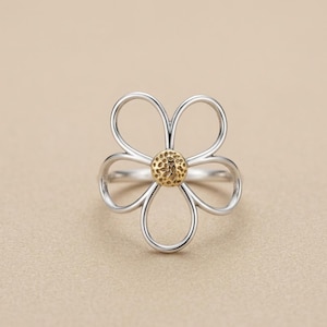 Puede incluir: Un anillo de plata que presenta un gran diseño de flor calada con cinco pétalos lisos y redondeados. El centro de la flor es un acento circular dorado texturizado con una superficie irregular y picada que contrasta con la banda de plata pulida.