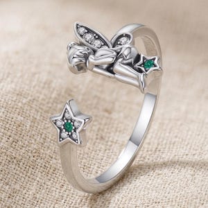 Puede incluir: Anillo plateado con diseño de hada. El hada está adornada con piedras preciosas claras y verdes. Dos detalles en forma de estrella, también con piedras preciosas, están en el anillo. El anillo tiene un diseño abierto.