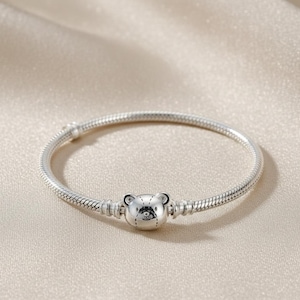 Puede incluir: Una pulsera de plata con un charm de oso. El charm es una cabeza de oso detallada con rasgos grabados. La pulsera tiene un diseño de cadena de serpiente y un cierre seguro. La joya se exhibe sobre una superficie neutra y texturizada.