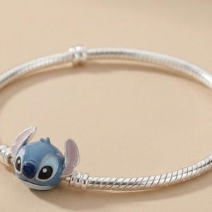 Peut inclure: Un bracelet en chaîne serpent argentée avec un charm représentant le personnage Stitch. Le charm est en émail bleu et rose avec des détails noirs et blancs, illustrant la tête de la créature. Le bracelet présente une finition polie et lisse.