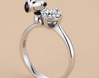 May include: A silver ring featuring a Snoopy figure holding a large, clear gemstone. The ring has a polished finish and the inscription "S925" is visible on the band. The Snoopy figure has black ears and a black nose.
