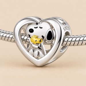 May include: A silver heart-shaped charm bead featuring a white Snoopy character holding a yellow Woodstock figure. The charm is threaded onto a silver snake chain bracelet. The side of the heart is engraved with the text S925, indicating sterling silver.